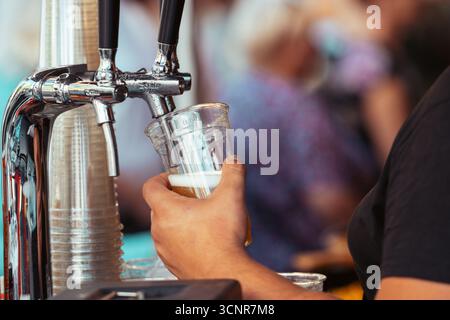 Primo piano di un barista che versa birra alla spina fresca in una tazza di plastica durante un festival all'aperto. Una lager fresca e schiumosa viene servita da una spina Foto Stock