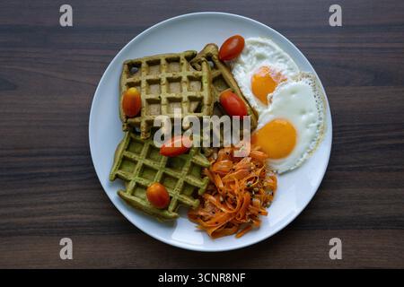 Vista dall'alto del piatto salutare della colazione con waffle verdi, uova fritte e verdure su un tavolo di legno, concetto di brunch, nutrizione. Foto Stock