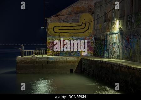 Zona di pesca notturna ad Almada in Portogallo con pareti illuminate di graffiti e un'atmosfera sul lungomare vicino a Lisbona Foto Stock