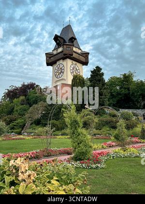 L'iconica Uhrturm (Torre dell'Orologio) sulla collina di Schlossberg si erge alto sopra i vivaci giardini, sotto un cielo estivo nel cuore storico di Graz. Foto Stock