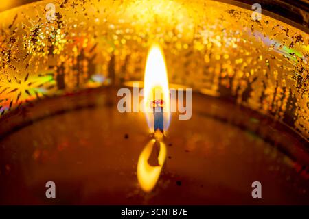 Una singola e vivace fiamma di candele illumina un contenitore decorativo dorato, creando una calda luce riflettente sulla cera fusa. Foto Stock