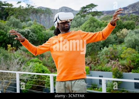 Uomo afroamericano che indossa una felpa arancione con visore VR bianco sul balcone con vista sulle colline Foto Stock