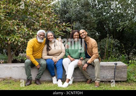 Famiglie diverse sedute su una panchina di legno in giardino e inclinate verso il treppiede della fotocamera Foto Stock