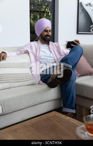 Uomo indiano che indossa turbante, camicia che scorre sullo smartphone sul divano con tazza di tè in vetro Foto Stock