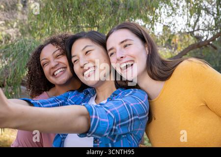 Diverse amiche si appoggiano e sorridono mentre scattano selfie all'aperto in un parco con smartphone Foto Stock