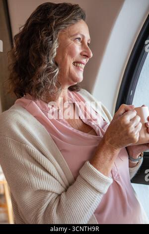 Donna anziana che indossa una camicetta e un cardigan mentre tiene in mano una tazza in ceramica bianca tramite una finestra curva Foto Stock