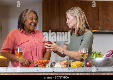 Diversi amici anziani in piedi nell'isola di cucina in marmo, affettare cetrioli e tenere il vino. Senior, amicizia, collaborazione, stile di vita sano, Mo Foto Stock