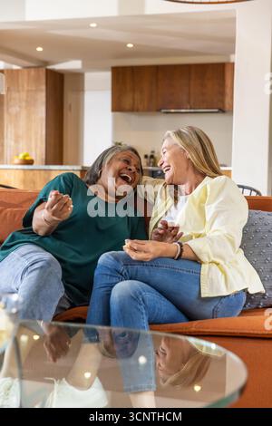 Diverse amiche anziane che ridono e condividono popcorn sul divano nel soggiorno open space Foto Stock