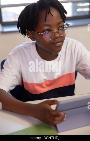 Studentessa afroamericana seduta alla scrivania in classe con occhiali da vista e tablet in mano Foto Stock
