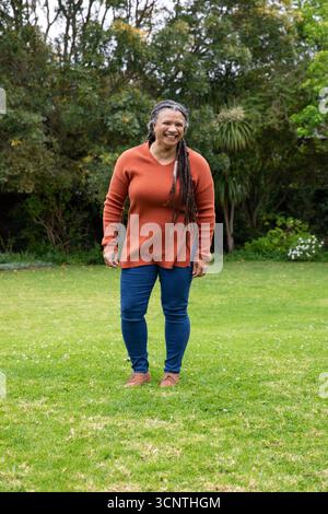 Donna afroamericana anziana in piedi mentre sorride sul prato del giardino indossando un maglione ruggine e jeans Foto Stock