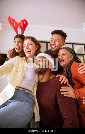 Diversi amici con la fascia da corna e il cappello di babbo natale che scattano selfie in salotto con lo smartphone Foto Stock