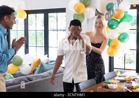 Diversi amici guidano un uomo afroamericano che copre gli occhi a casa con palloncini e striscioni Foto Stock