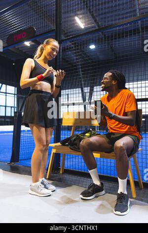 Diversi partner di allenamento che riposano sulla panchina con una borsa di palle da tennis, utilizzando smartphone sul campo Foto Stock