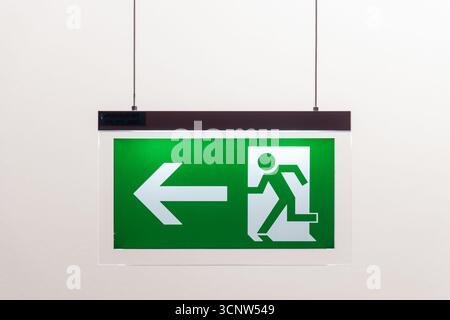 Segnale di uscita di emergenza illuminato con l'icona di una persona e una freccia rivolta verso sinistra appesa al soffitto Foto Stock