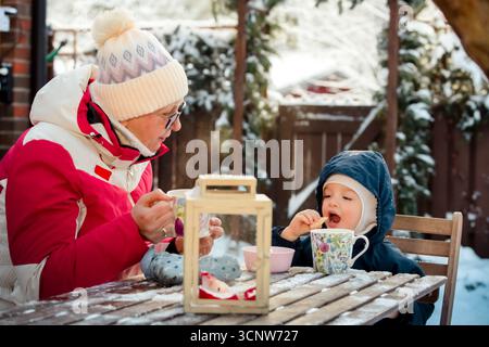 Nonna e bambino si godono cioccolata calda e biscotti all'aperto in una giornata invernale innevata, intimo momento in famiglia con un'atmosfera festosa e stagionale. Foto Stock
