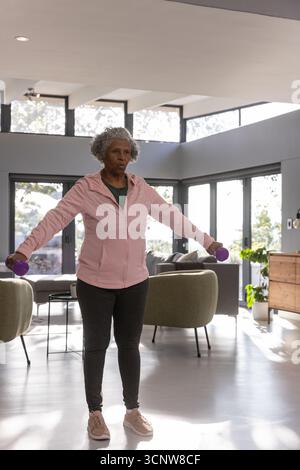 Donna afroamericana anziana che indossa abiti sportivi, solleva manubri viola in salotto Foto Stock