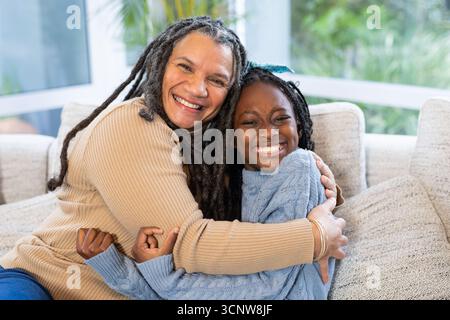 La nonna anziana e la nipote si abbracciano sul divano vicino alle finestre con cuscini nella stanza del sole Foto Stock