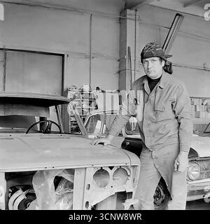 Un maestro carrozziere posa per un ritratto accanto al telaio smantellato di una VAZ 'Zhiguli' in un'officina di riparazione a Sloviansk, RSS Ucraina, durante gli anni '1980 Con uno sguardo serio e diretto, l'abile artigiano si erge nel suo ambiente di lavoro autentico, circondato da auto in fase di revisione, tra cui un Moskvitch-412 sullo sfondo. Questa fotografia in bianco e nero è una potente rappresentazione dell'artigianato e del lavoro industriale del tardo periodo sovietico Foto Stock