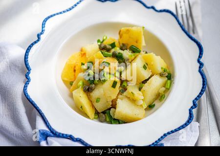 Insalata di patate greche guarnita con capperi, servita in un elegante piatto a coste blu. Questo delizioso piatto combina ingredienti freschi e salati. Foto Stock