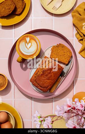 Vista dall'alto di una torta di carote fatta in casa servita con caffè su un vassoio rosa su uno sfondo con piastrelle rosa. Gli oggetti circostanti includono piatti, uova e fiori Foto Stock