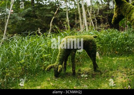 Una scultura di cervo ricoperta di muschio sorge tra una lussureggiante vegetazione in un parco forestale. I dettagli intricati si fondono perfettamente con l'ambiente naturale circostante, Foto Stock