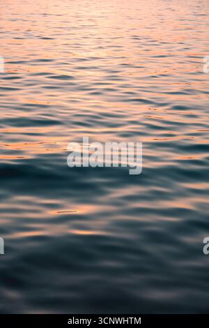 Una serena vista macro delle dolci onde del mare, illuminata dal caldo bagliore di un tramonto. Le increspature creano un motivo rilassante, riflettendo le sfumature delicate del rosa Foto Stock