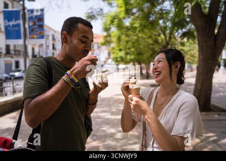 Una coppia gioiosa condivide un momento delizioso gustando i coni di gelato in una vivace piazza della città durante una giornata di sole, catturando l'essenza del divertimento estivo a. Foto Stock