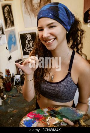 Una giovane artista con un velo blu si trova nel suo studio d'arte con una tavolozza colorata. Vari dipinti e schizzi adornano le pareti Foto Stock
