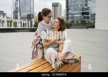Una figlia adolescente abbraccia amorevolmente sua madre da dietro mentre si siedono su una panchina di legno in una moderna piazza urbana. Lo skyline della città e le fontane Foto Stock