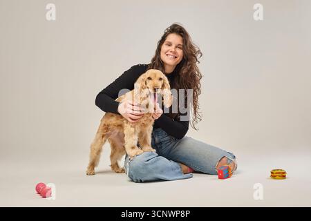 Donna sorridente seduta sul pavimento con il suo adorabile cucciolo spaniel cocker, circondato da giocattoli colorati. Un momento allegro che mostra il legame tra di loro Foto Stock