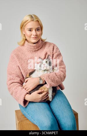 Una donna con i capelli biondi si siede comodamente tenendo in mano un gatto in un accogliente ambiente da studio. La donna indossa un morbido maglione rosa e il gatto ha un aspetto attento Foto Stock