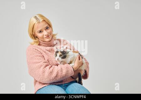 Una donna allegra con i capelli biondi, vestita con un comodo maglione, tiene amorevolmente il suo gatto in uno studio. L'atmosfera trasuda calore e compagnia tra di noi Foto Stock
