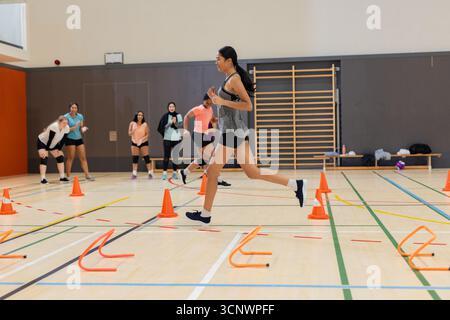 Atleta che salta su coni arancioni e ostacoli in palestra con i compagni in attesa Foto Stock