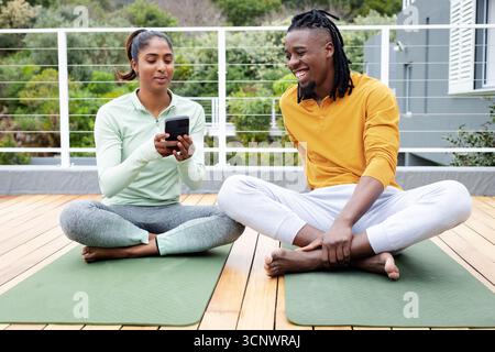 Coppia indiana e afroamericana in abbigliamento sportivo seduta su tappetini da yoga in condivisione smartphone sulla terrazza Foto Stock