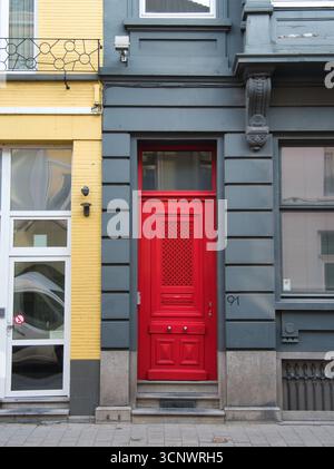 Vibrant Red Door su una facciata grigia - Una porta rossa brillante e audace con un motivo geometrico e delle maniglie delle portiere lucide si distingue contro una faca grigia a contrasto Foto Stock