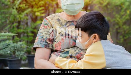 Vista laterale ritratto del nipote abbracciando sua nonna e indossando una maschera medica in un parco. Concetto di famiglia sano. Foto Stock