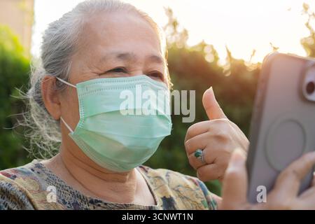Anziana donna asiatica anziana che indossa la maschera usando il suo cellulare per parlare con la sua famiglia su sfondo naturale. Foto Stock