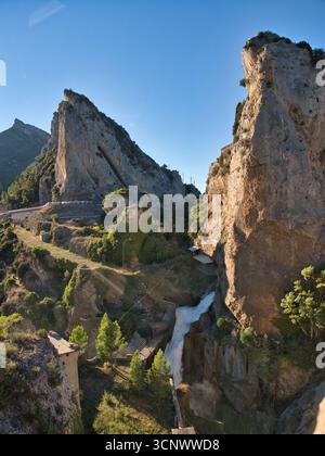 Erosione tra due scogliere di montagna - Una potente cascata scende lungo un'insenatura rocciosa tra due torreggianti scogliere di montagna, con una strada tortuosa che tortuosa Foto Stock