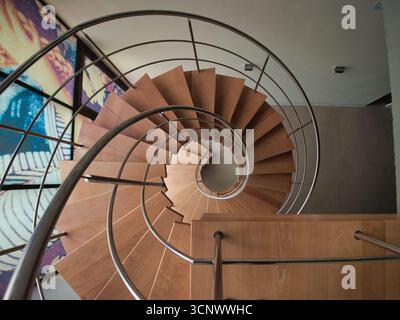 Vista dall'alto verso il basso della scala a spirale - Un'immagine ravvicinata ad angolo alto di una moderna scala a chiocciola con gradini in legno chiaro e un elegante corrimano in metallo, crea Foto Stock