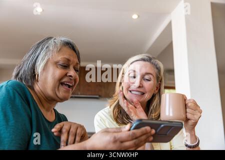 Diverse amiche anziane che tengono in mano smartphone e tazza, chiacchierano al bancone della cucina Foto Stock