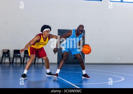 Atleti afro-americani maschi che dribblano pallacanestro arancione e difendono in campo in palestra Foto Stock