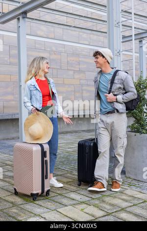 Coppia in piedi su marciapiede piastrellato sotto la tettoia di vetro all'ingresso dell'edificio, con valigie rotanti Foto Stock
