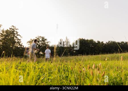 Un padre e il suo giovane figlio trascorrono del tempo all'aperto in un lussureggiante campo verde durante un caldo pomeriggio. Camminano insieme, godendo della natura e di ogni altra parte Foto Stock