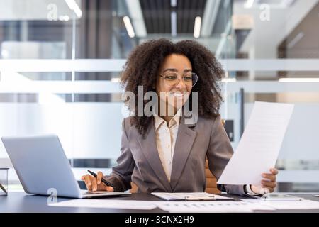 Una giovane donna d'affari afro-americana sorridente lavora in ufficio, seduto a una scrivania con un computer portatile e in possesso di documenti e bollette. Foto Stock