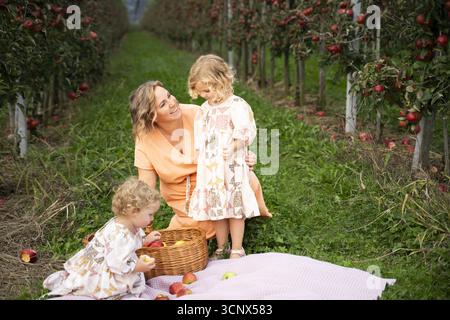 Una famiglia allegra gode di una giornata di sole in un frutteto di mele, circondato da lussureggianti alberi di mele. I bambini raccolgono le mele mentre la loro madre guarda avanti Foto Stock