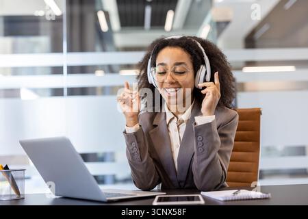 Giovane donna afroamericana seduta in ufficio con le cuffie alla scrivania. ascolta musica e balli. Foto Stock