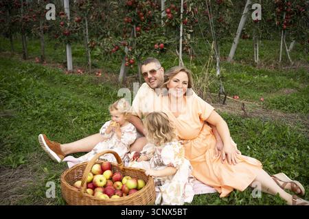 Una famiglia allegra gode di una giornata di sole seduto sull'erba in un meleto, circondato da mele mature. I bambini giocano e assaggiano frutta fresca da a. Foto Stock