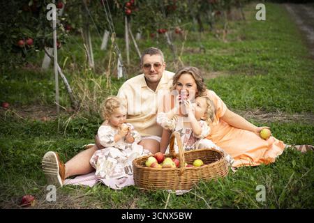 Una famiglia gioiosa ama trascorrere una giornata nel meleto, seduto sull'erba con un cesto di mele. I genitori e i loro figli condividono le risate e la fresca a Foto Stock