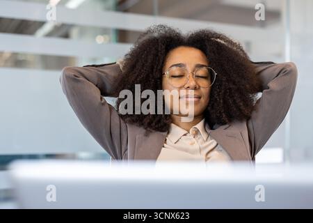 Foto ravvicinata di una giovane donna d'affari afroamericana sorridente seduta a una scrivania in ufficio, che chiude gli occhi e riposa durante una pausa. Foto Stock