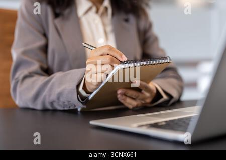 Foto ravvicinata delle mani di una giovane donna afroamericana con un abito da lavoro e una camicia. Seduto alla scrivania di fronte a un computer portatile, tenendo una penna e prendendo appunti in un notebook. Foto Stock